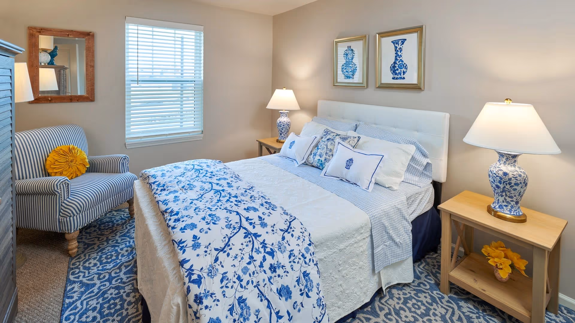 A cozy bedroom with a neatly made bed featuring white and blue floral bedding and multiple pillows. On either side of the bed are wooden nightstands with matching blue and white patterned lamps. A striped armchair with a yellow flower-shaped cushion sits near a window with white blinds. The room has beige walls, two framed blue and white vase prints above the bed, and a blue patterned rug on the floor.