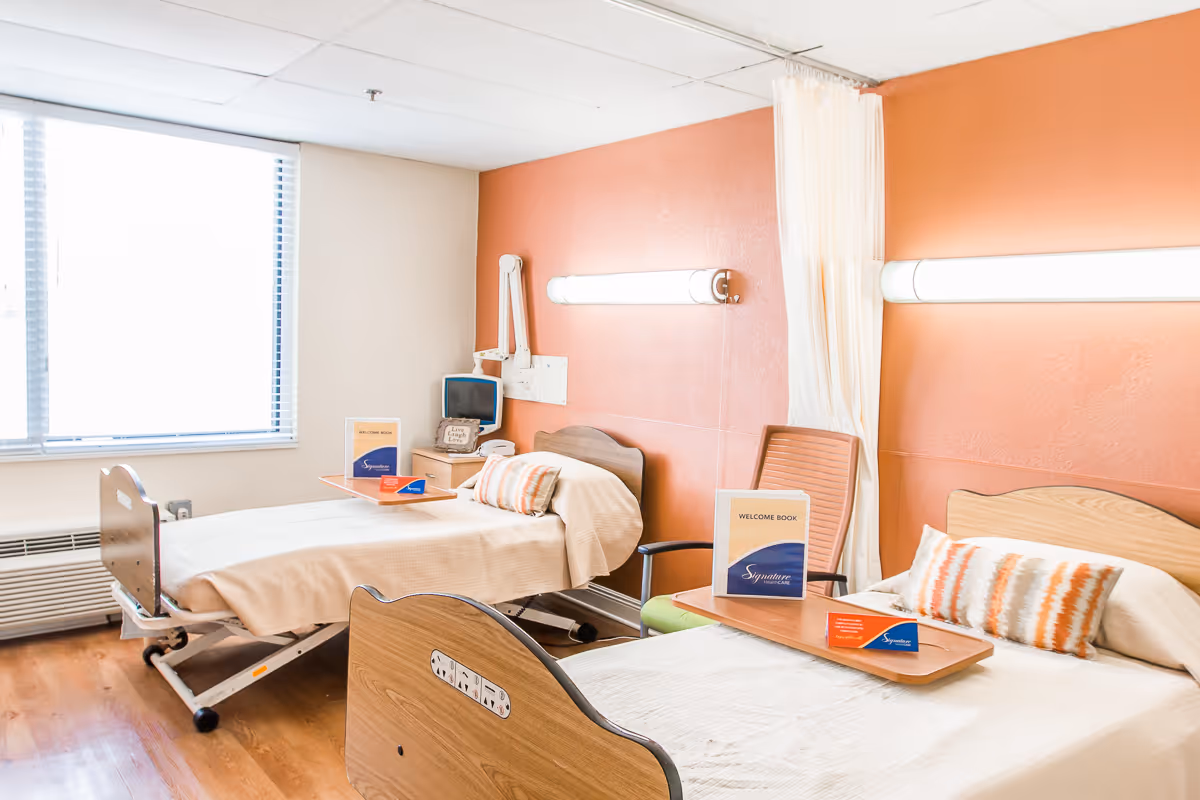 Two hospital-style beds in a bright senior care room with an orange accent wall, bedside trays, pillows, and a large window.