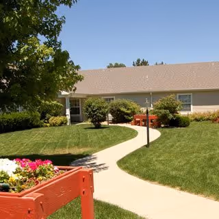 A paved walkway curves through a well-maintained lawn with green grass and bushes, leading to the entrance of a single-story building with beige siding and a brown roof. There is a red wooden planter box with colorful flowers in the foreground on the left side, and a tree providing shade on the left.