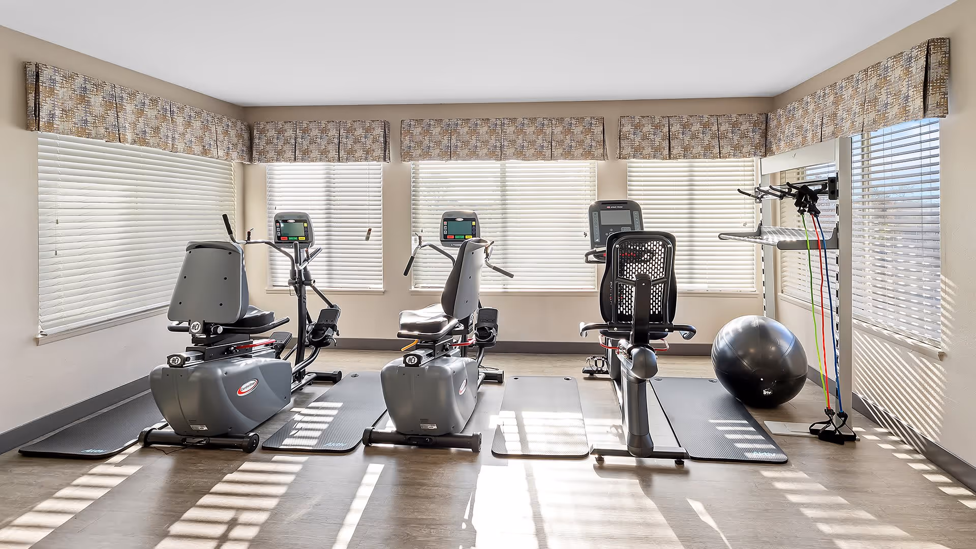 A bright exercise room with large windows covered by blinds and valances. The room contains three recumbent exercise bikes placed on individual mats, a large black exercise ball, and a rack holding resistance bands. The floor is wood-style, and sunlight streams through the windows creating shadows on the floor.