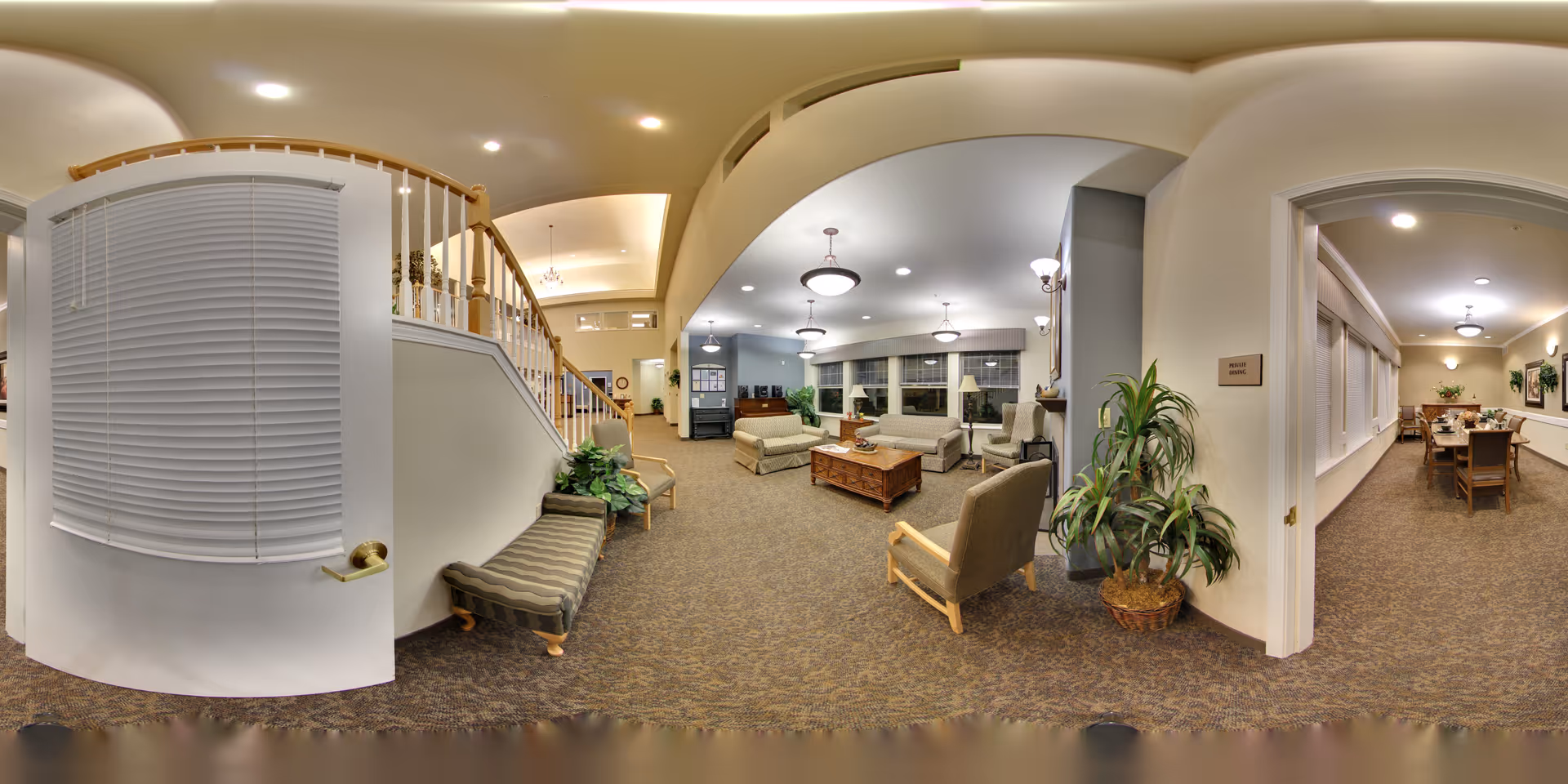 A spacious senior living facility common area with comfortable seating including sofas and armchairs arranged around a wooden coffee table. The room is well-lit with ceiling lights and has large windows with blinds. To the right, there is an open doorway leading to a dining room with a long table and chairs. The area features carpeted floors, potted plants, and a staircase with wooden railings.