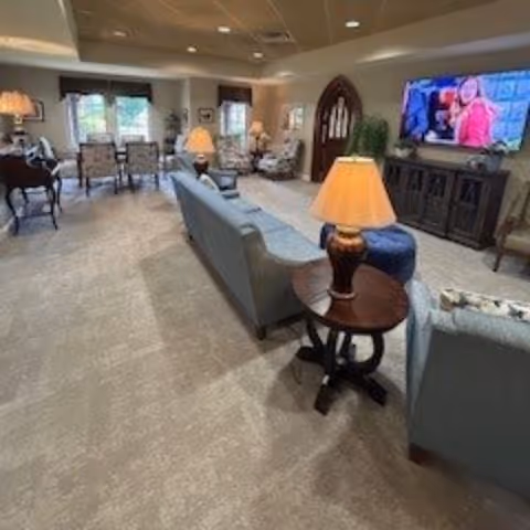 A spacious living room area with multiple seating options including sofas and armchairs arranged around side tables with lamps. A large flat-screen TV is mounted on the wall above a dark wooden cabinet. The room has carpeted flooring, several windows with curtains, and soft lighting creating a cozy atmosphere.