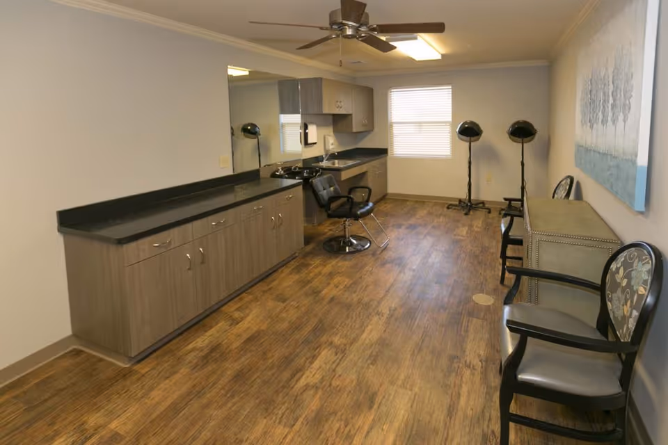 Interior salon-style room with cabinets, a styling chair and hooded hair dryers, plus seating along the right wall.