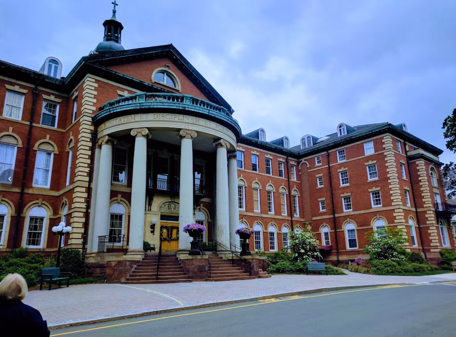 Front exterior view of a large, historic brick building with tall white columns at the entrance, a cross on top of the roof, and well-maintained landscaping including bushes and flowers. A person with blonde hair is walking on the sidewalk in front of the building.