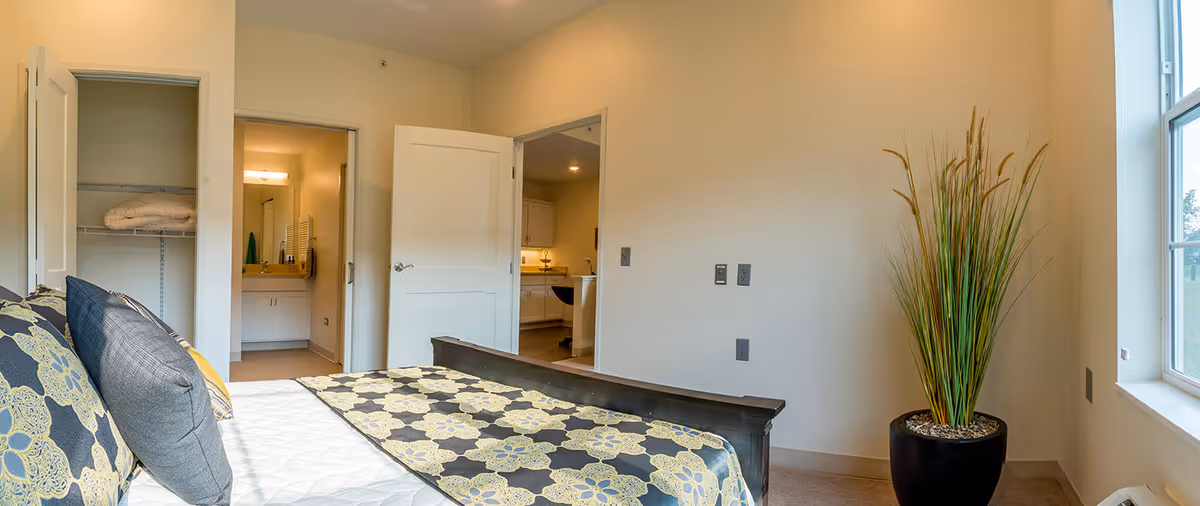 A bedroom with a bed featuring a patterned bedspread and pillows. There is an open closet door with folded towels inside, an open door leading to a bathroom with a sink and mirror, and another open door leading to a kitchen area. A tall potted plant is placed near a window letting in natural light.