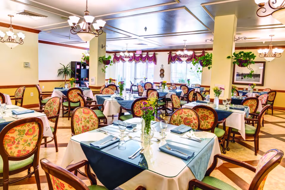 Dining room with neatly set tables, floral centerpieces, and patterned upholstered chairs in a senior living facility.