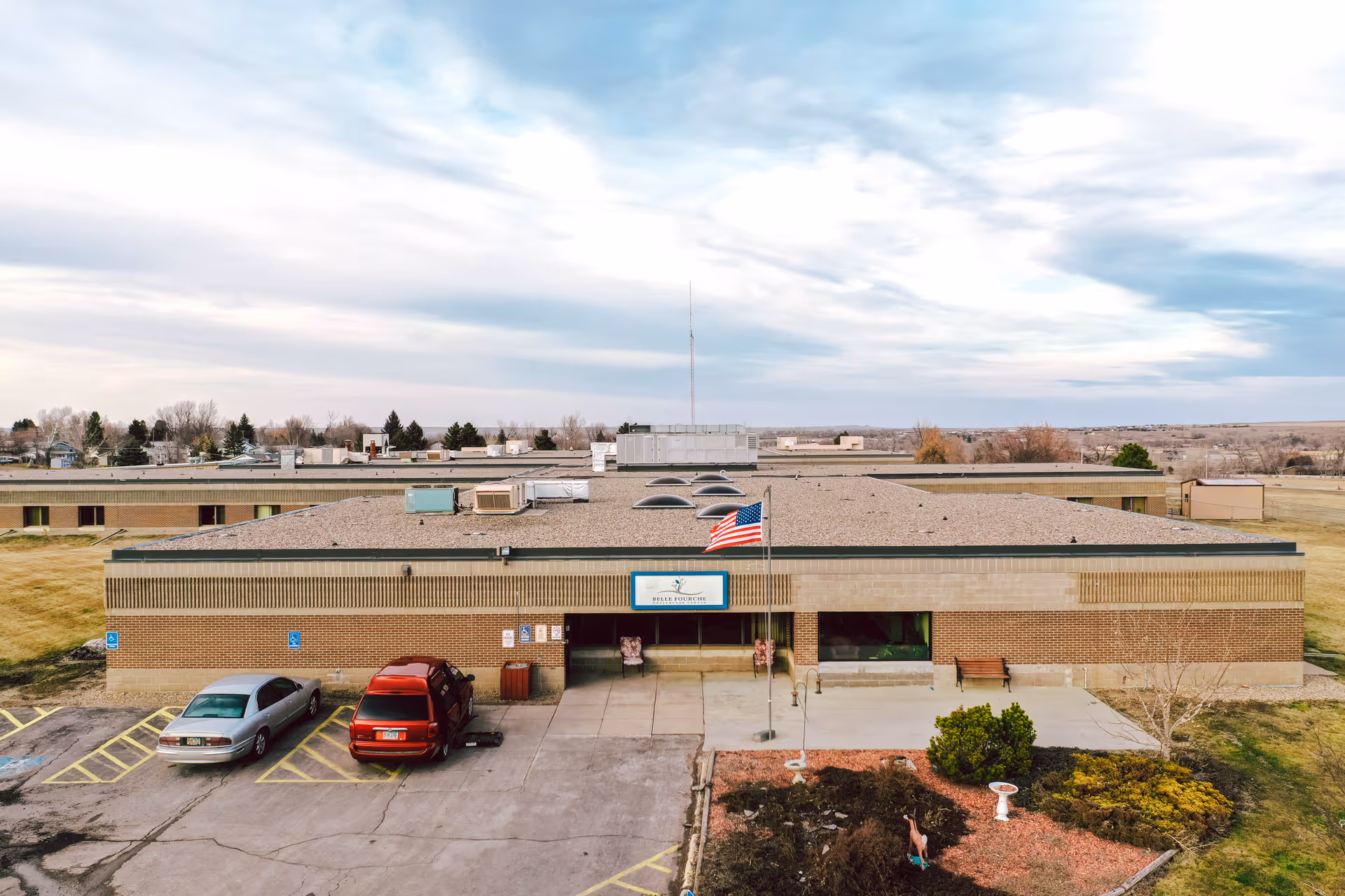 Front exterior view of a single-story brick building with a flat roof, an American flag on a flagpole in front, two cars parked in designated spots, and a small landscaped area with bushes and a birdbath. The sky is partly cloudy.