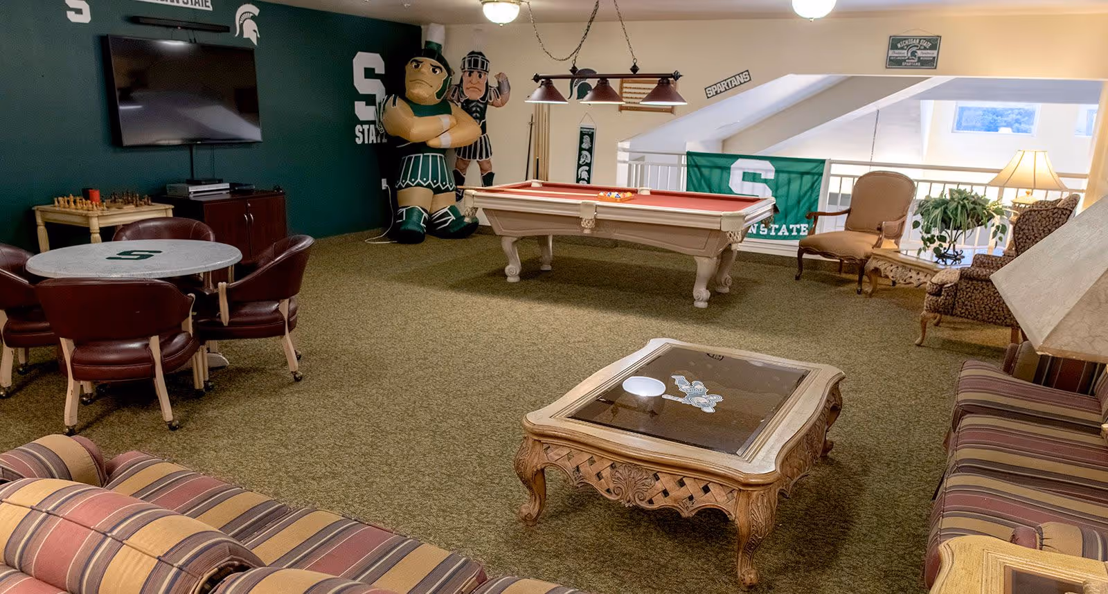 A recreational room decorated with Michigan State University Spartans memorabilia, including a large inflatable Spartan mascot, a pool table, a round table with four chairs, a chess set, a TV mounted on a green wall, and comfortable seating with a striped couch and armchairs. The room has carpeted flooring and a hanging light fixture above the pool table.