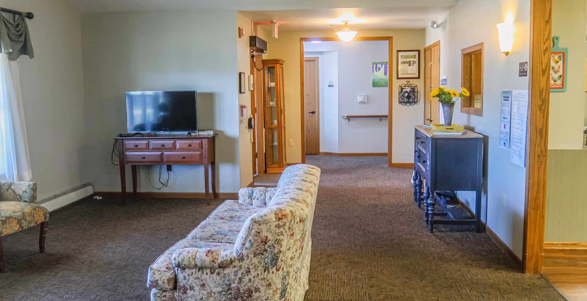A cozy senior living facility common area with floral upholstered sofas and chairs, a flat-screen TV on a wooden console table, and a hallway leading to several doors. The walls are decorated with framed pictures and a vase with yellow flowers on a side table.
