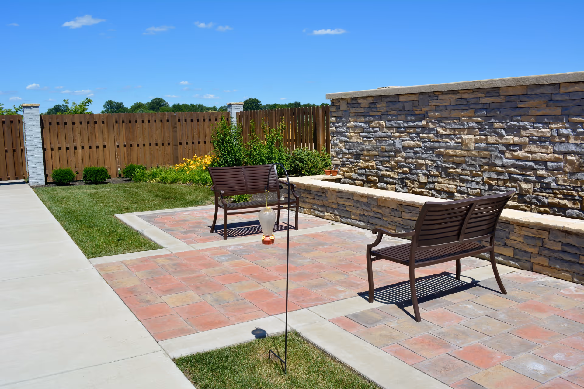 Outdoor patio area with two brown metal benches on a tiled surface, a stone wall with a ledge, a wooden fence, green grass, and some bushes and flowers under a clear blue sky.
