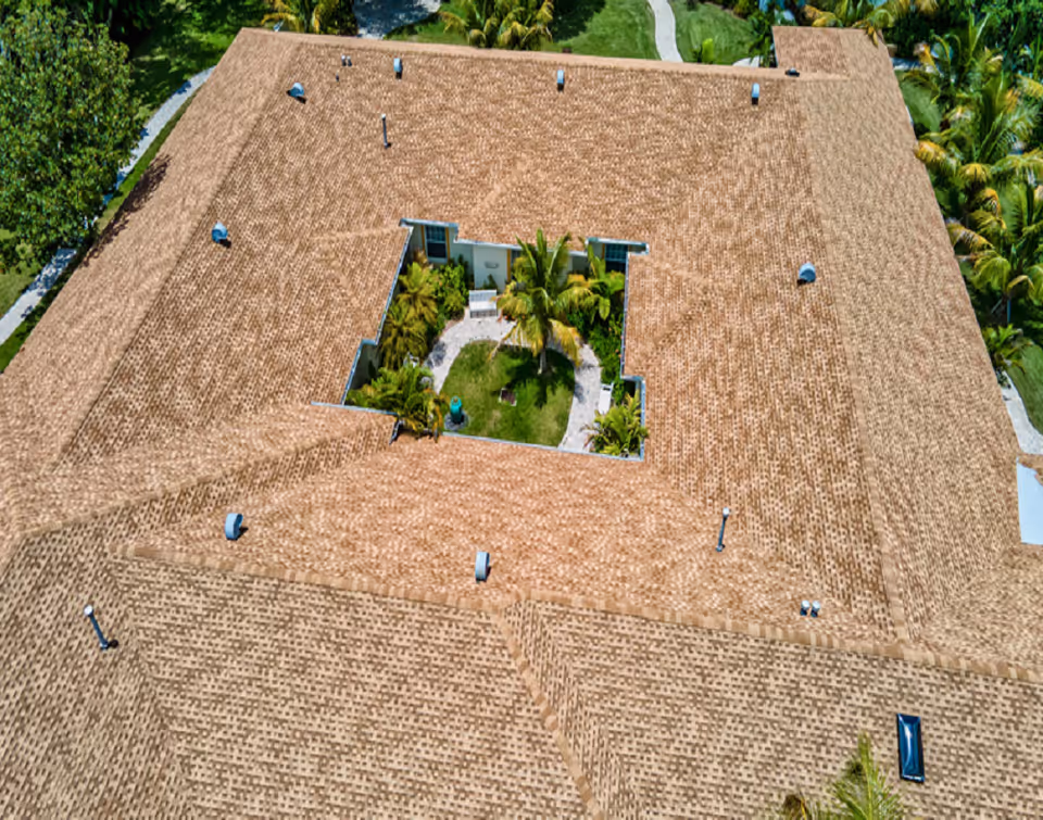 Aerial view of a building with a large, brown shingle roof surrounding a central courtyard garden with palm trees, green grass, and a walking path.