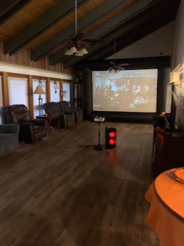 Interior of a room with wooden flooring and a high ceiling with exposed beams. There are several recliner chairs along the left wall next to windows with blinds. A large projector screen is set up at the far end of the room, displaying a blurry image. In front of the screen, there is a projector on a stand and a speaker with red lights. Ceiling fans with lights hang from the ceiling. A round table with an orange tablecloth is partially visible on the right side.