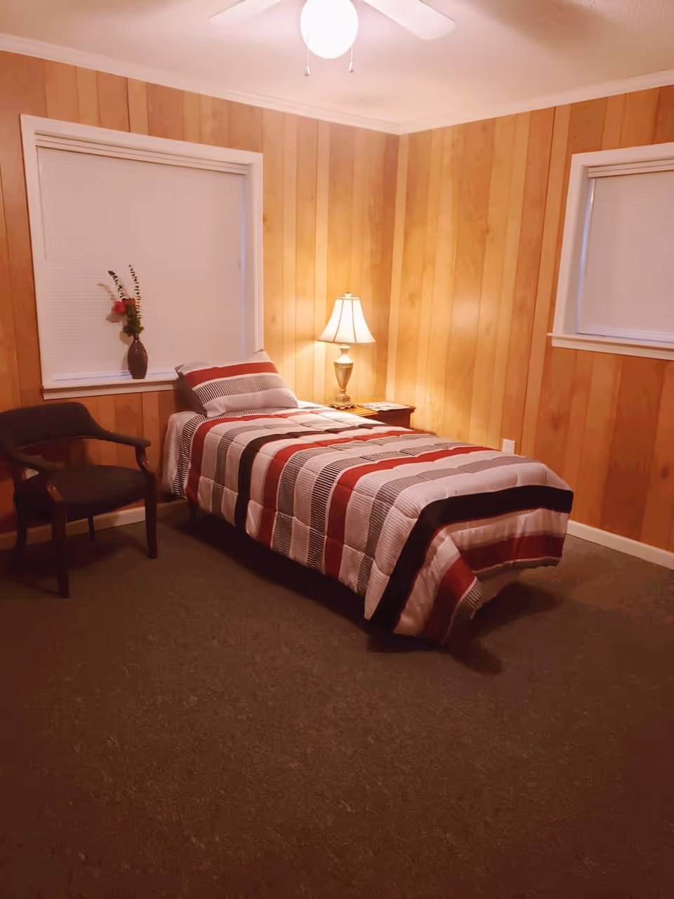 Single bed with striped bedding in a wood-paneled bedroom with a bedside lamp, chair, and two windows.