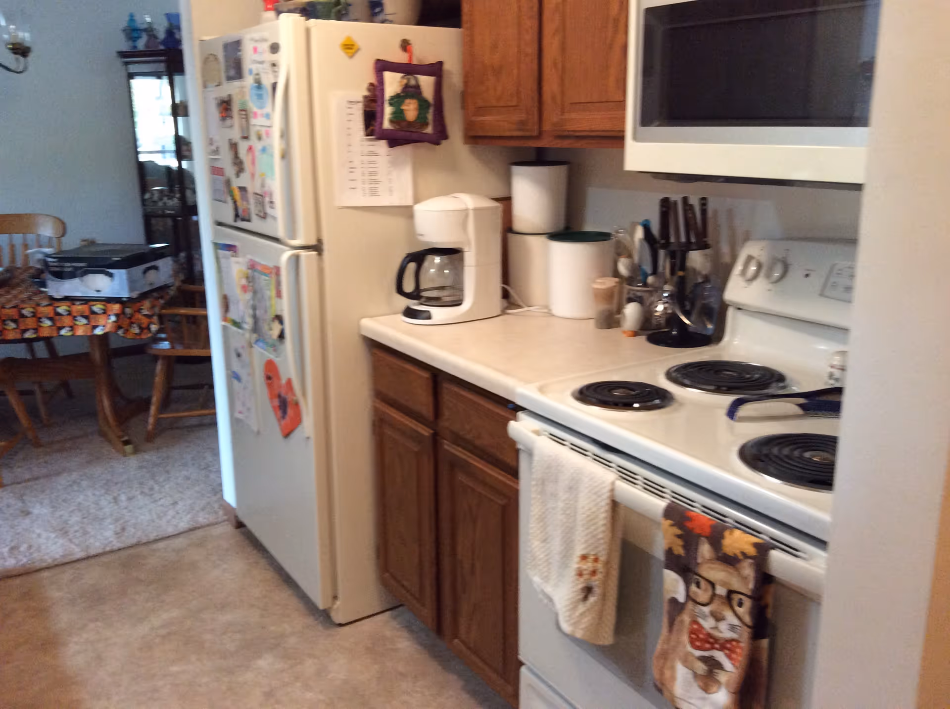 A kitchen area with a white refrigerator covered in magnets and papers, a white coffee maker on the counter, wooden cabinets, a white electric stove with oven, and a microwave above the stove. In the background, there is a dining area with a wooden table and chairs, and a tablecloth with a Halloween-themed pattern.