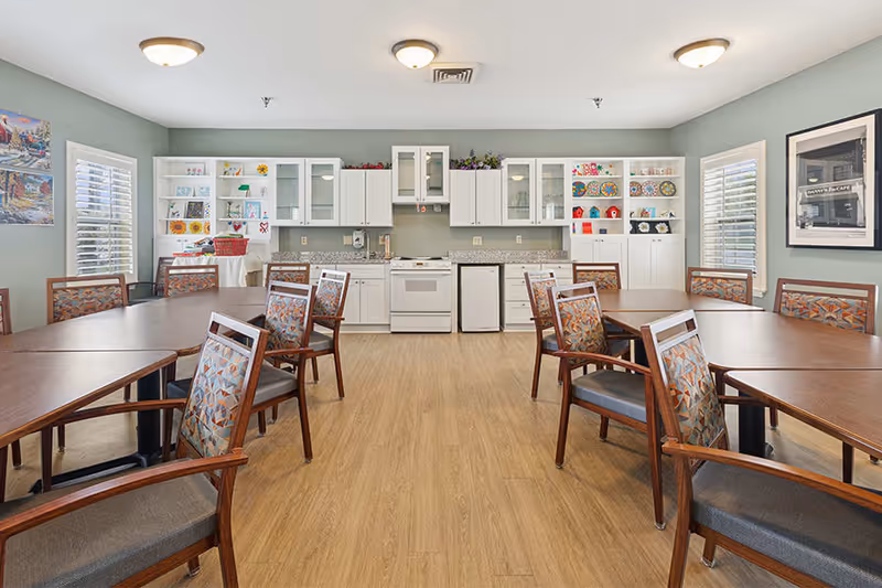 A bright dining room with multiple wooden tables and chairs arranged neatly. The back wall features a kitchen area with white cabinets, a stove, a small refrigerator, and shelves displaying colorful plates and decorative items. The room has light green walls, wood flooring, and windows with white shutters allowing natural light to enter.