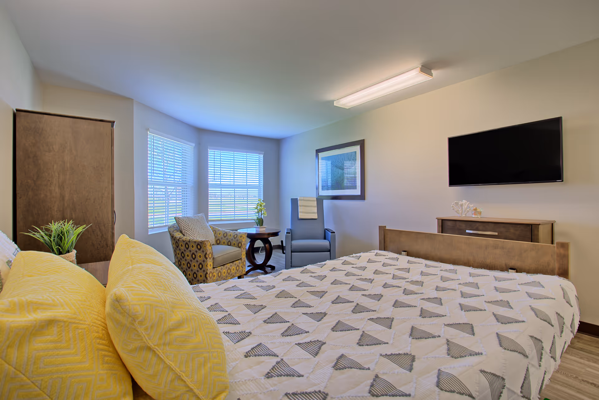 A bright bedroom with a large bed covered in a white and gray patterned quilt and yellow pillows. There is a wooden dresser with a flat-screen TV mounted above it on the wall. Two chairs and a small round table are positioned near two windows with white blinds, allowing natural light to fill the room. A framed picture hangs on the wall above one of the chairs.