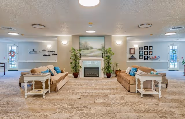 A spacious and well-lit common area with two beige sofas facing each other, each with blue and beige cushions. Between the sofas are white side tables with decorative items. In the center background is a fireplace flanked by two tall green plants and a large framed landscape painting above it. The room has a patterned carpet and white walls with wall-mounted lights and various framed pictures and decorations. Doors with glass panels are visible on both sides of the room.