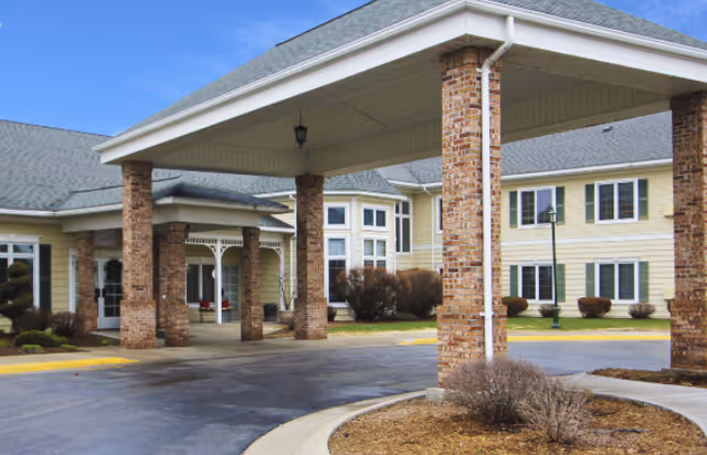 Exterior view of a senior living facility named LakeHouse Escanaba showing a covered entrance supported by brick columns, with a driveway and landscaped bushes in front of a two-story building with multiple windows.