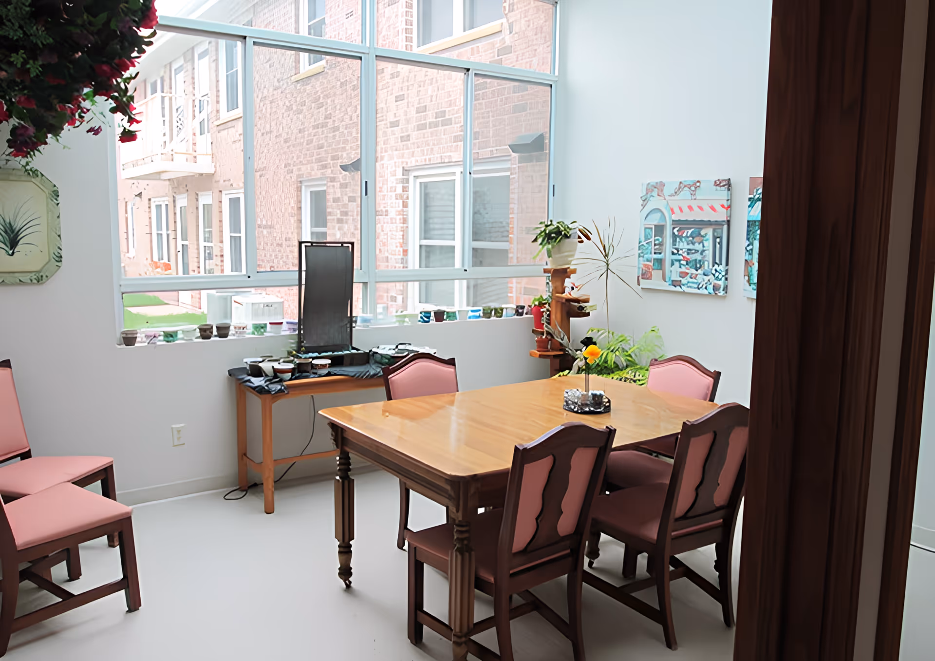 A bright dining room with a wooden table surrounded by six chairs with pink cushions. There is a large window showing a brick building outside. A small table near the window holds various small pots and a decorative water feature. The room has white walls with two colorful paintings and several plants, including a hanging plant and a potted plant on a wooden stand.