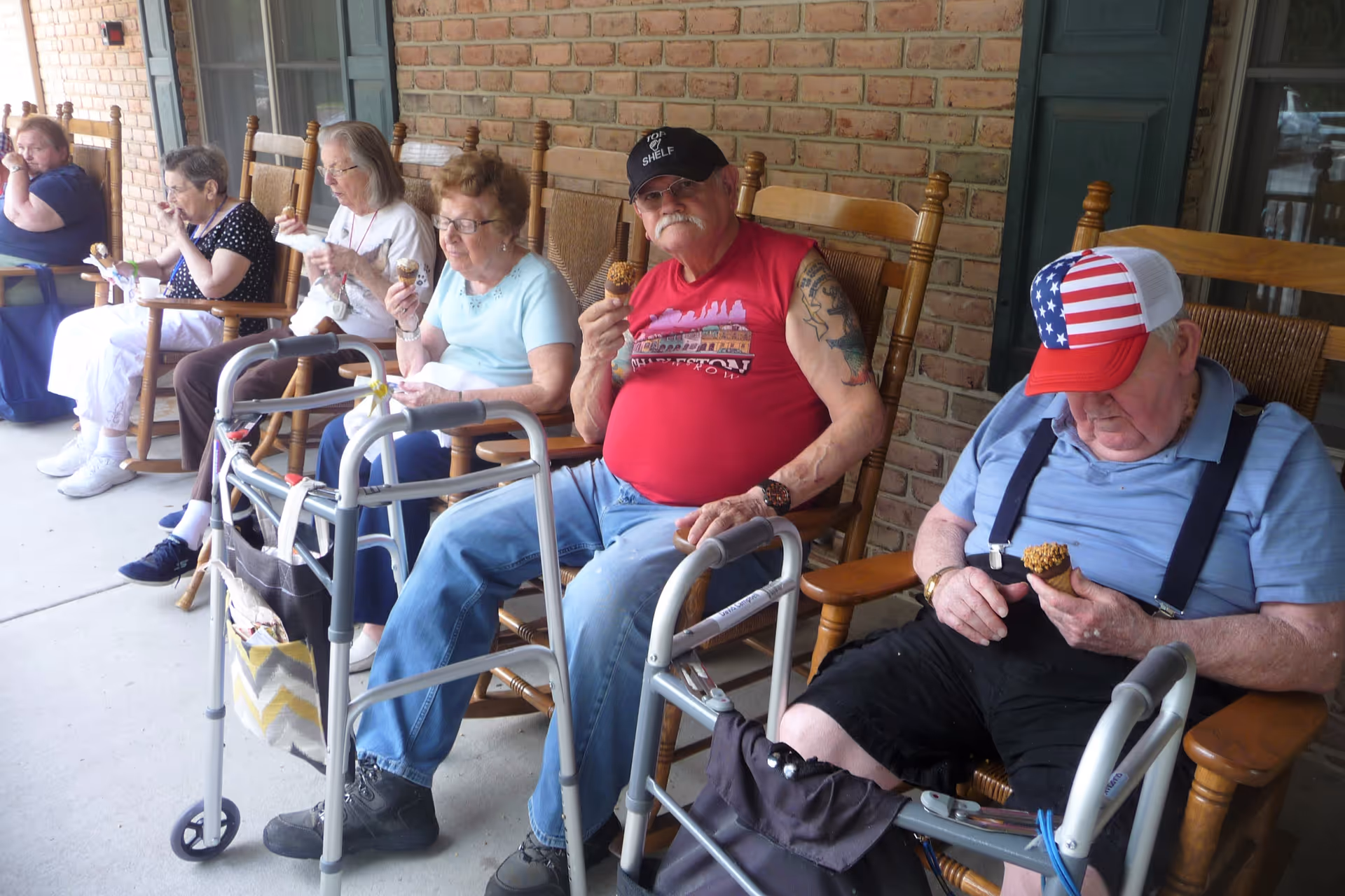 A group of elderly people sitting in wooden rocking chairs on a covered porch, eating ice cream cones. Two walkers are positioned in front of some of the chairs. The porch has a brick wall and a green door in the background.