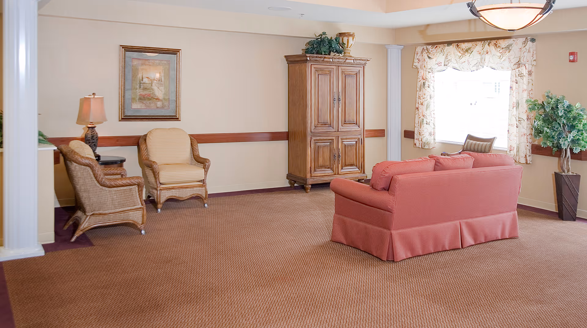 Spacious lounge with a red sofa, wicker armchairs, a wooden armoire, and a window with floral curtains.