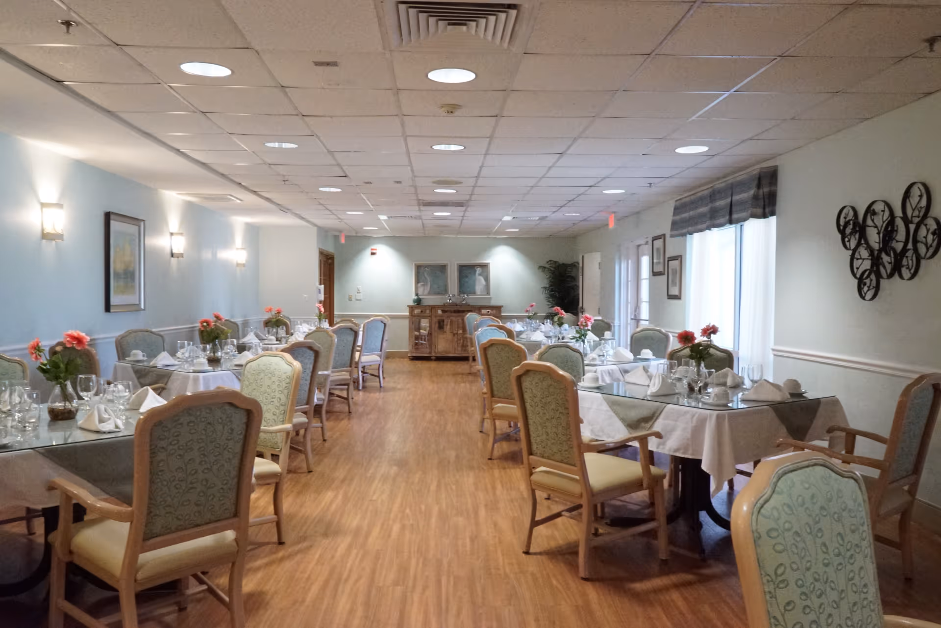A dining room with multiple tables set with white tablecloths, glassware, napkins, and small flower arrangements. The room has wooden flooring, light-colored walls, and several upholstered chairs around each table. Wall sconces and framed artwork decorate the walls, and there is a sideboard at the far end of the room.