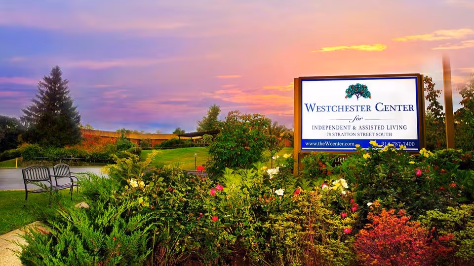 A landscaped garden area with various green shrubs and colorful flowers in front of a large sign for Westchester Center for Independent & Assisted Living at 78 Stratton Street South, with a sunset sky in the background and a bench on the left side.