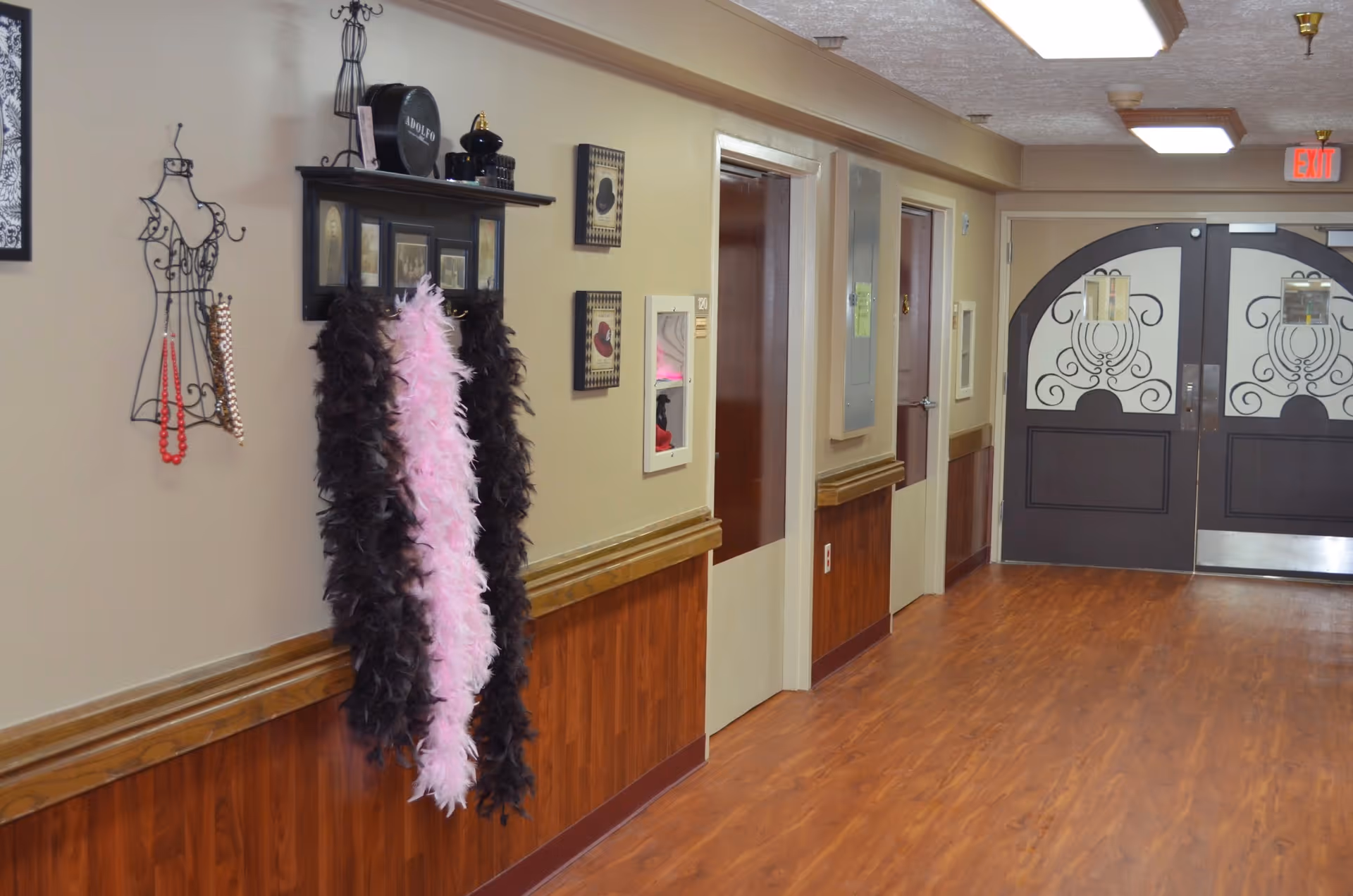 A hallway in a senior living facility with wood flooring and beige walls. On the left wall, there are decorative items including a wire mannequin with necklaces, a shelf with framed pictures and hats, and two feather boas hanging. The hallway has several doors and ends with double doors featuring decorative glass panels. Ceiling lights and an exit sign are visible.