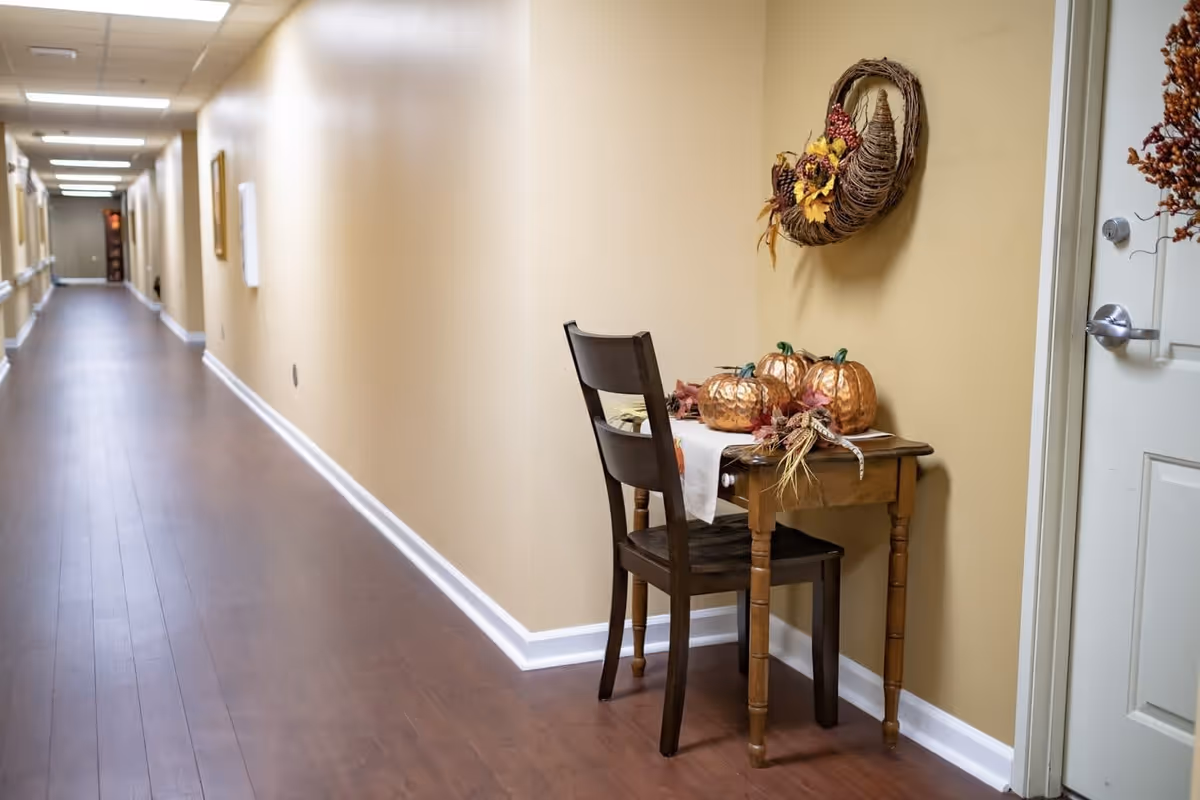 A long hallway with wooden flooring and beige walls in a senior living facility. On the right side, there is a small wooden table with a dark wooden chair. The table is decorated with three metallic pumpkins and autumn-themed decorations. A fall wreath hangs on the wall above the table. The hallway is well-lit with ceiling lights and has several doors and framed pictures along the walls.