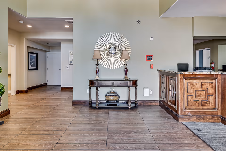 Interior view of a senior living facility lobby area with a wooden reception desk on the right, a decorative console table with two lamps and a circular mirror on the wall in the center, and a hallway leading to other rooms on the left. The floor is tiled with a wood-like pattern and the walls are painted light gray.
