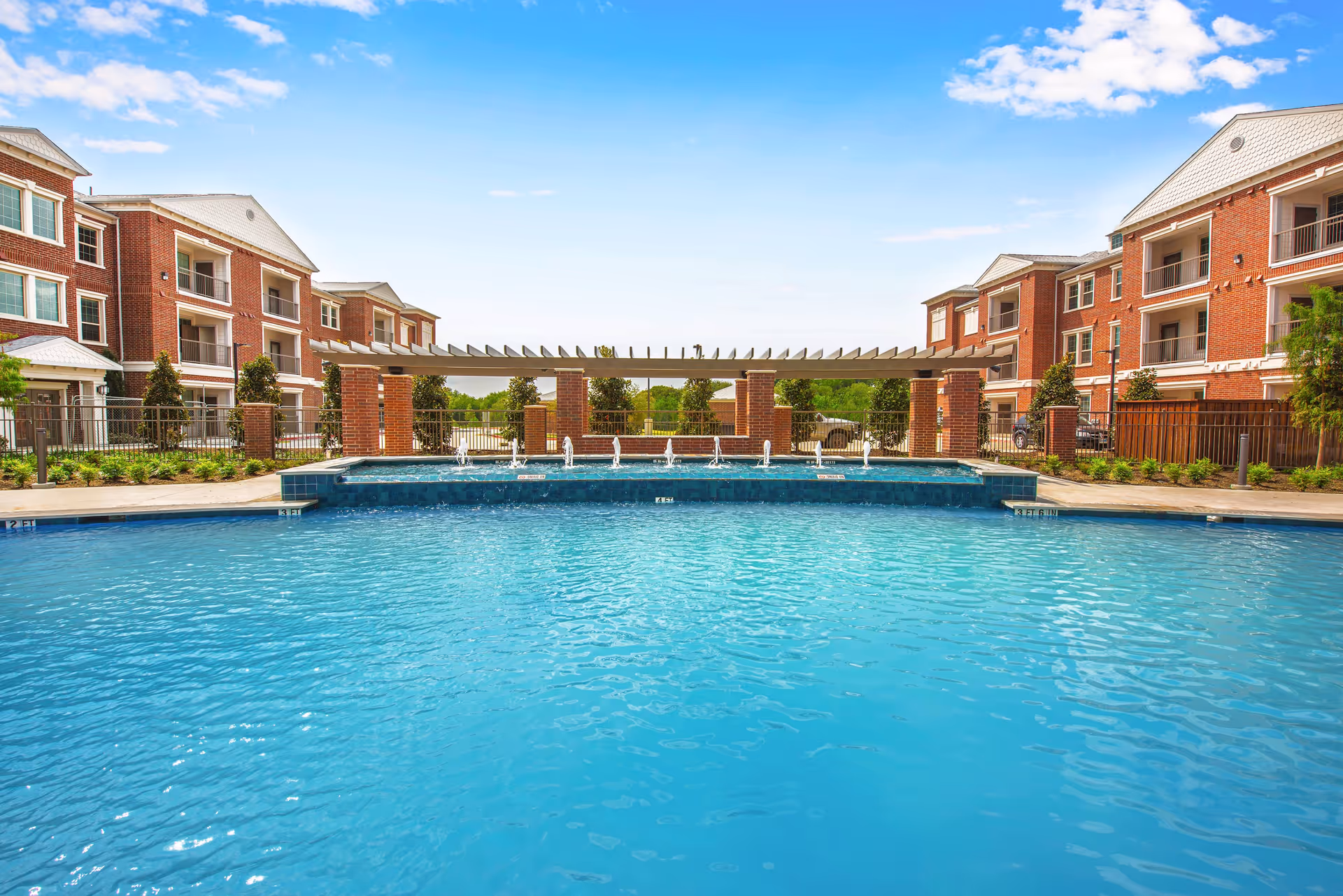 Outdoor swimming pool with water fountains in front of a pergola and surrounded by multi-story brick residential buildings under a blue sky with some clouds.