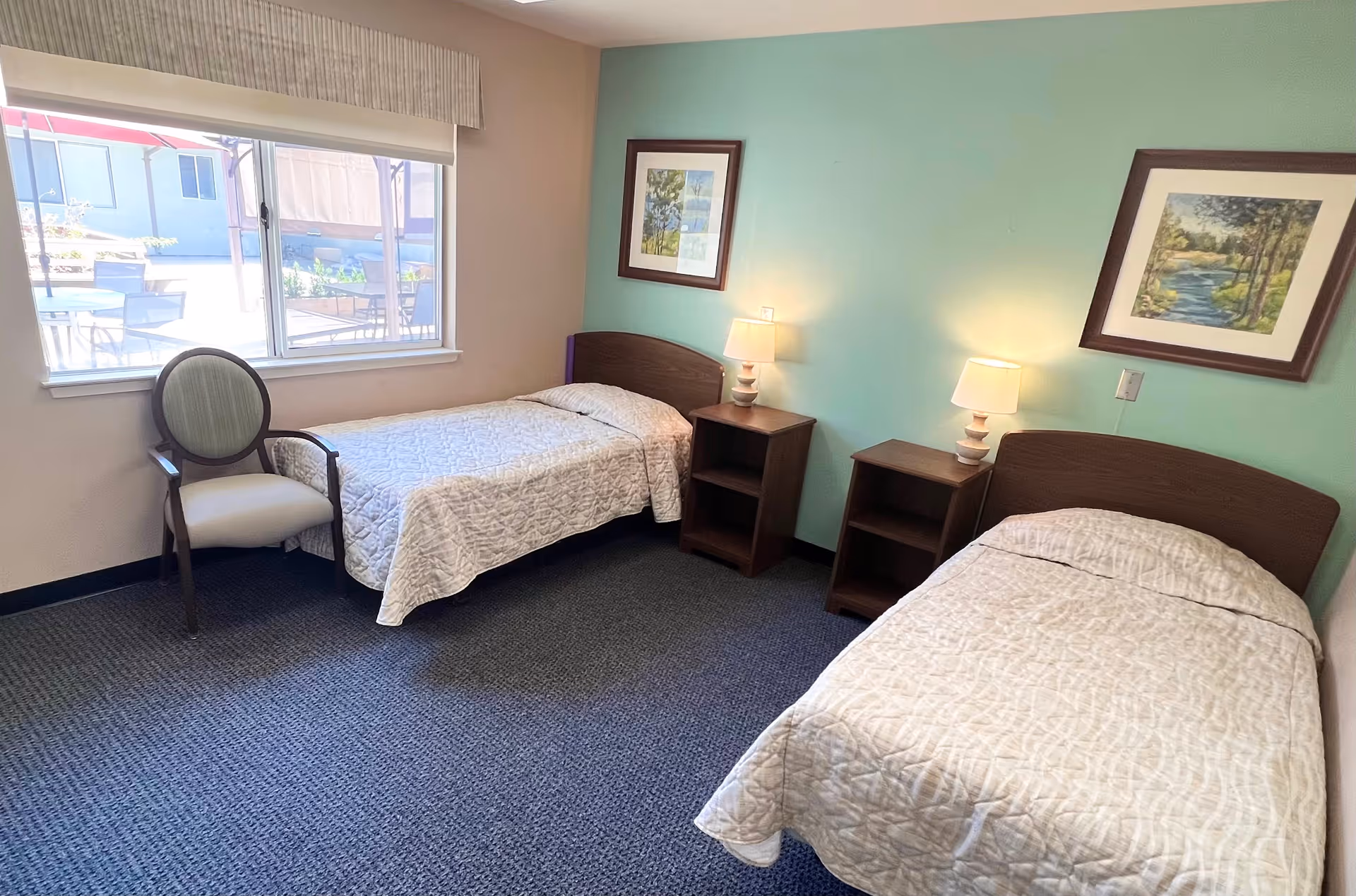 A simple bedroom with two single beds covered in light-colored quilts, each with a wooden headboard. Between the beds are two wooden nightstands, each with a lamp. There are two framed landscape paintings on the light green wall above the beds. A window on the left side lets in natural light, and a cushioned chair with wooden arms is placed near the window. The floor is carpeted in a dark color.