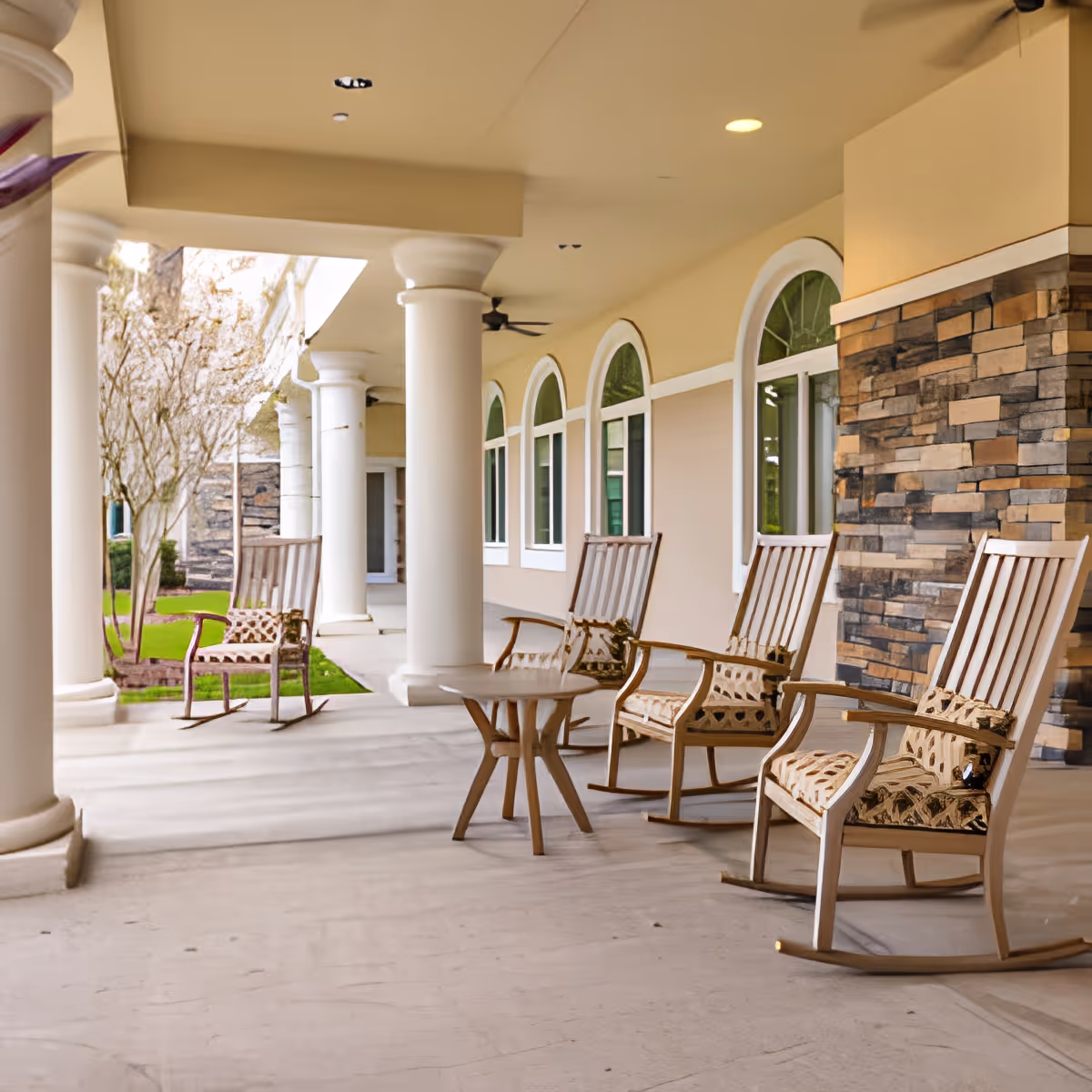 Covered outdoor patio area with several wooden rocking chairs with patterned cushions arranged around small round tables. The patio features large white columns and arched windows along the building wall, with a stone accent wall on one side and some greenery visible in the background.
