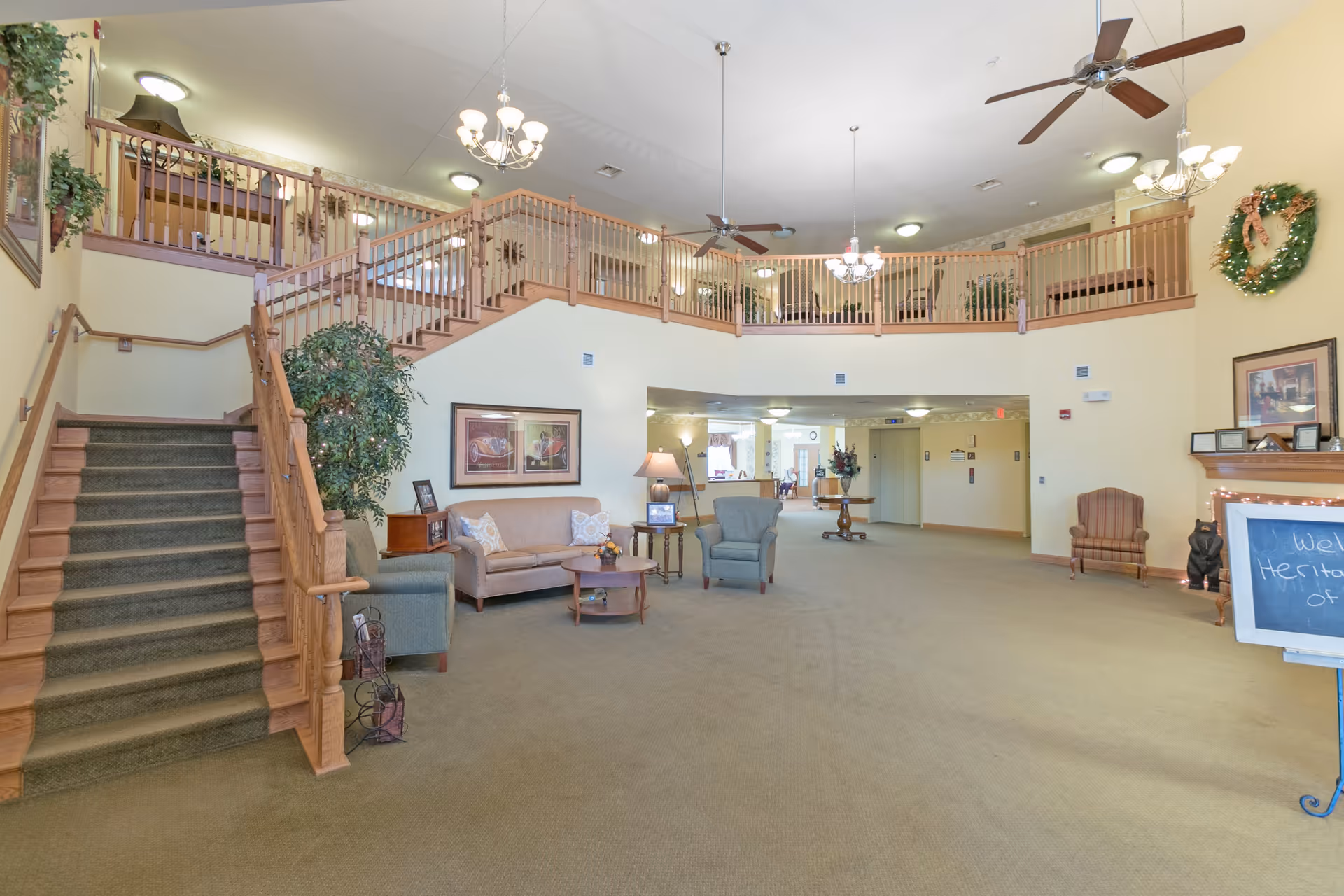 Spacious senior living facility lobby with a staircase on the left leading to an upper balcony. The area features comfortable seating including a sofa and armchairs, decorative plants, framed artwork on the walls, ceiling fans, chandeliers, and a wreath on the right wall. A chalkboard sign is visible on the right side near a fireplace mantel.