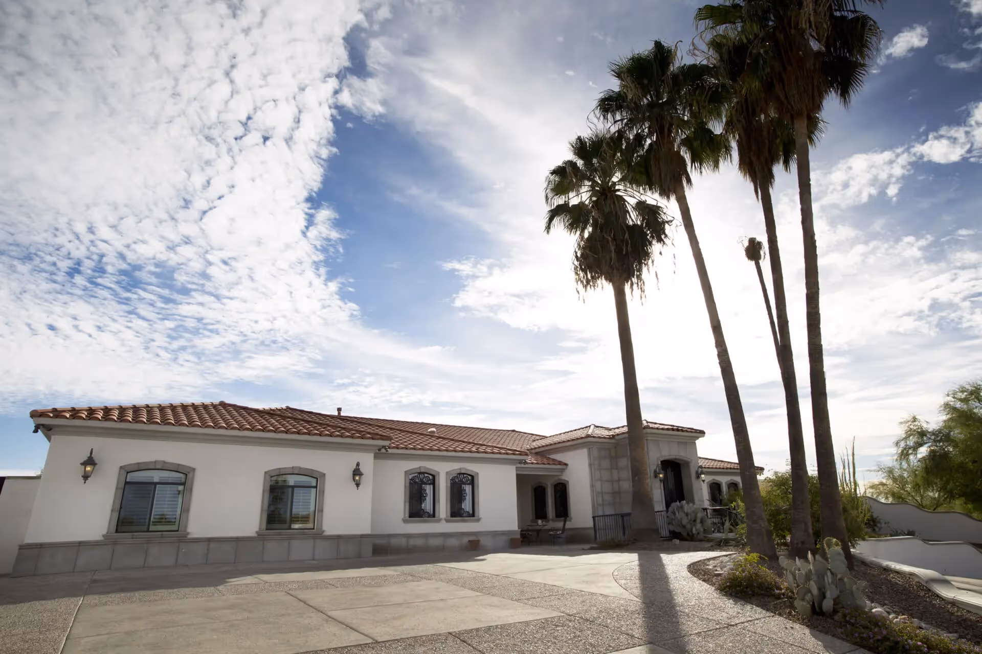Single-story Mediterranean-style building with a red tile roof, tall palm trees and a driveway under a partly cloudy sky.