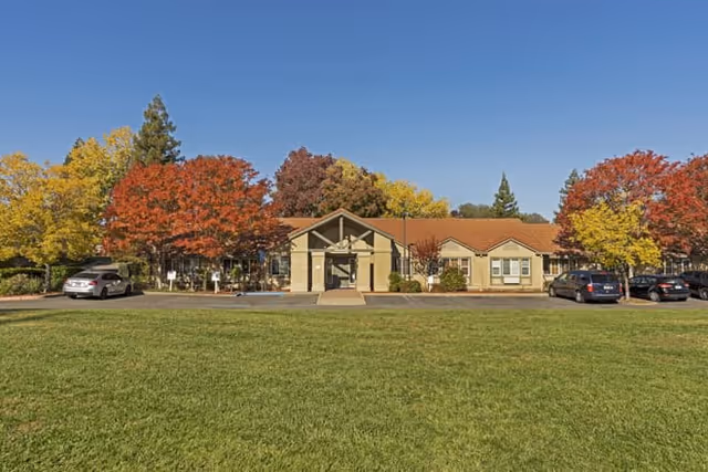 Single-story building with a brown roof and beige walls surrounded by trees with autumn foliage, a green lawn in the foreground, and several parked cars on either side of the building under a clear blue sky.