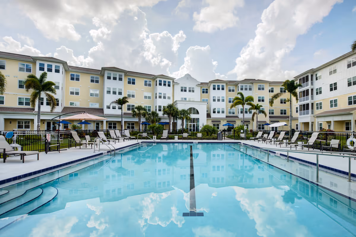 Outdoor swimming pool with clear blue water surrounded by lounge chairs and umbrellas, set in front of a multi-story senior living facility building with palm trees and a partly cloudy sky.