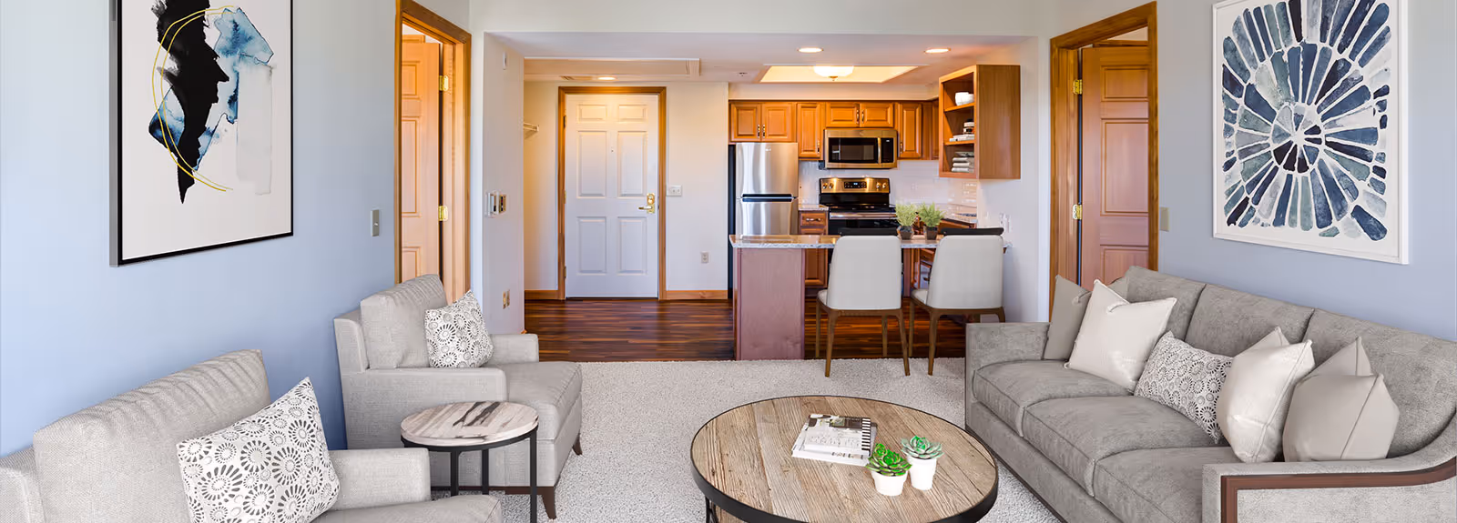 A cozy living room with light gray sofas and armchairs, a round wooden coffee table with books and small plants, and two abstract paintings on the light blue walls. The room opens into a kitchen area with wooden cabinets, stainless steel appliances, and a small dining counter with two chairs. The entrance door is visible in the background.