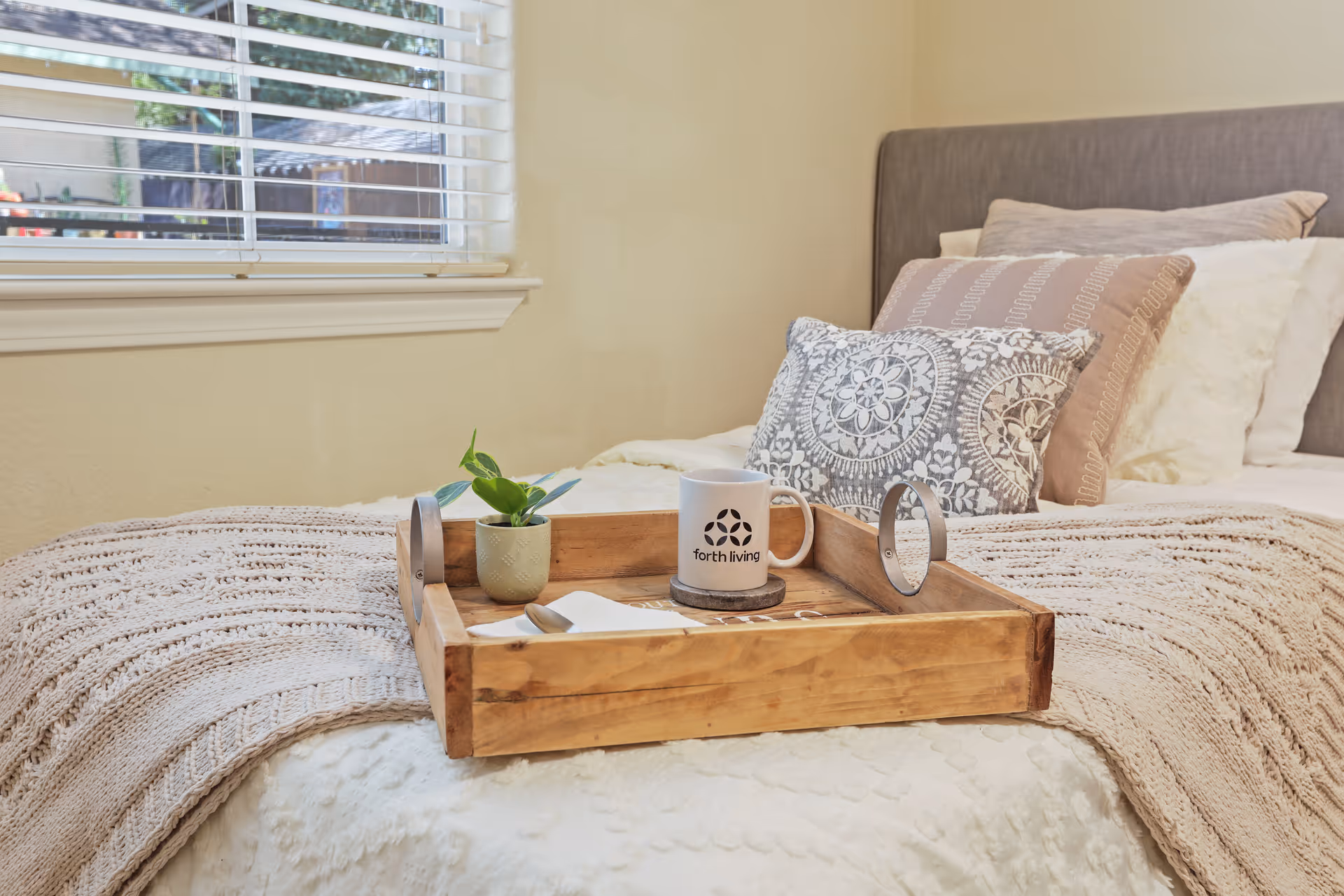 A cozy bedroom scene with a neatly made bed featuring a knitted beige blanket and decorative pillows. On the bed is a wooden tray holding a small potted plant and a white mug with the text 'forth living'. A window with white blinds is visible in the background.