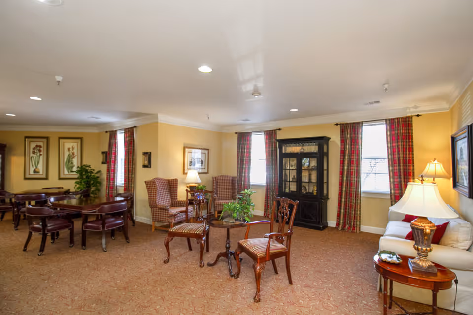 A bright communal living room with upholstered armchairs, wooden chairs and tables, a china cabinet, and plaid curtains.