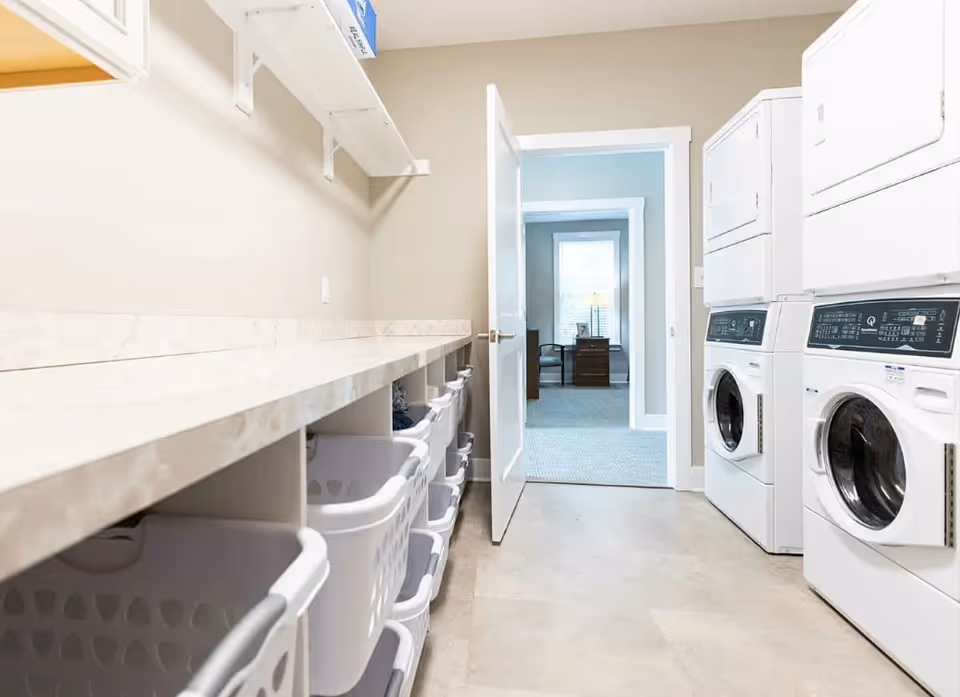 Laundry room with stacked washers and dryers, a long folding counter and multiple laundry baskets with an open door leading to another room.