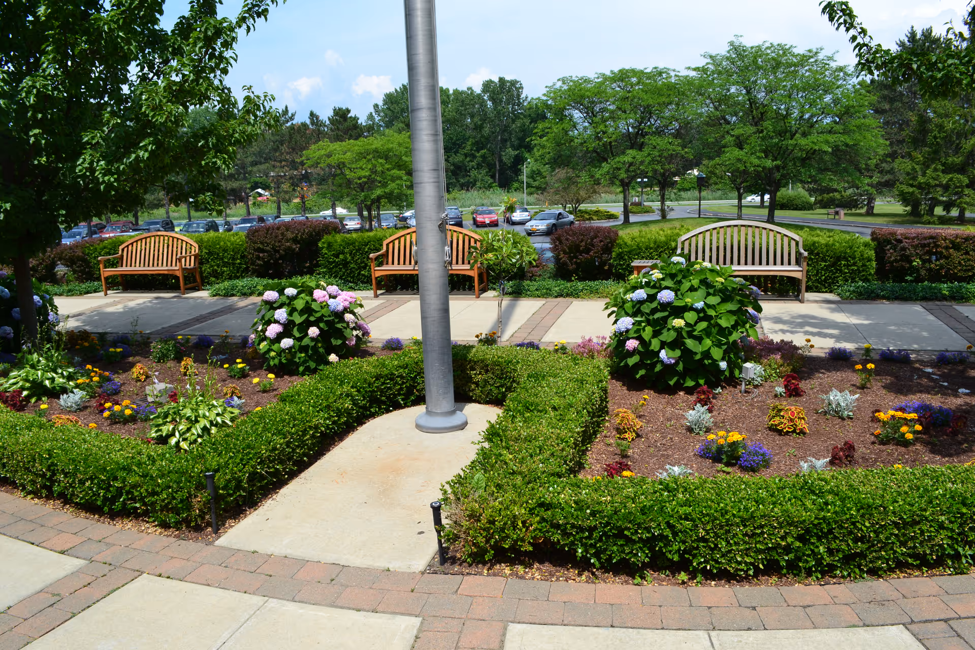 A well-maintained outdoor garden area with colorful flowers and neatly trimmed bushes surrounding a flagpole. There are three wooden benches on a paved walkway, with trees and parked cars visible in the background under a partly cloudy sky.