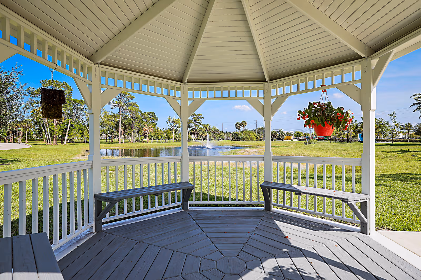 View from inside a white wooden gazebo with built-in benches, overlooking a grassy area with a pond and a fountain in the distance under a clear blue sky. There are hanging flower pots with red flowers on the gazebo.