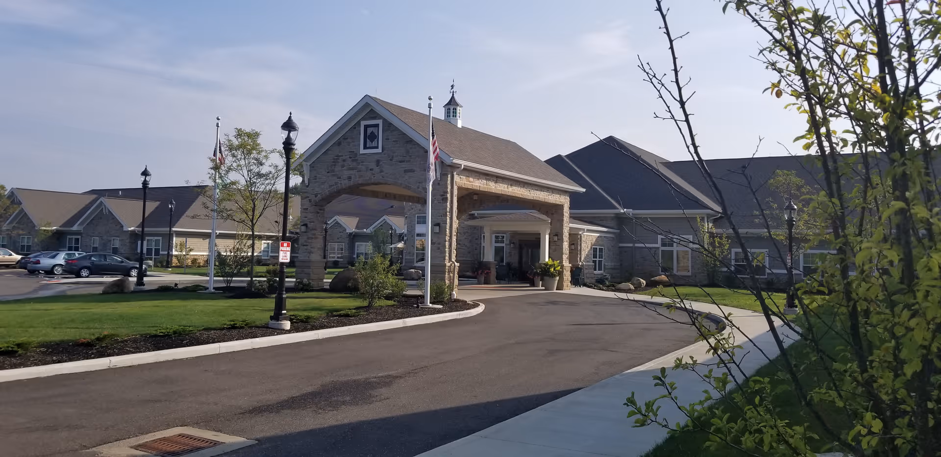 Exterior view of The Gables of Green senior living facility showing a large stone entrance with a covered driveway, surrounded by well-maintained lawns, trees, and a parking area with several cars.