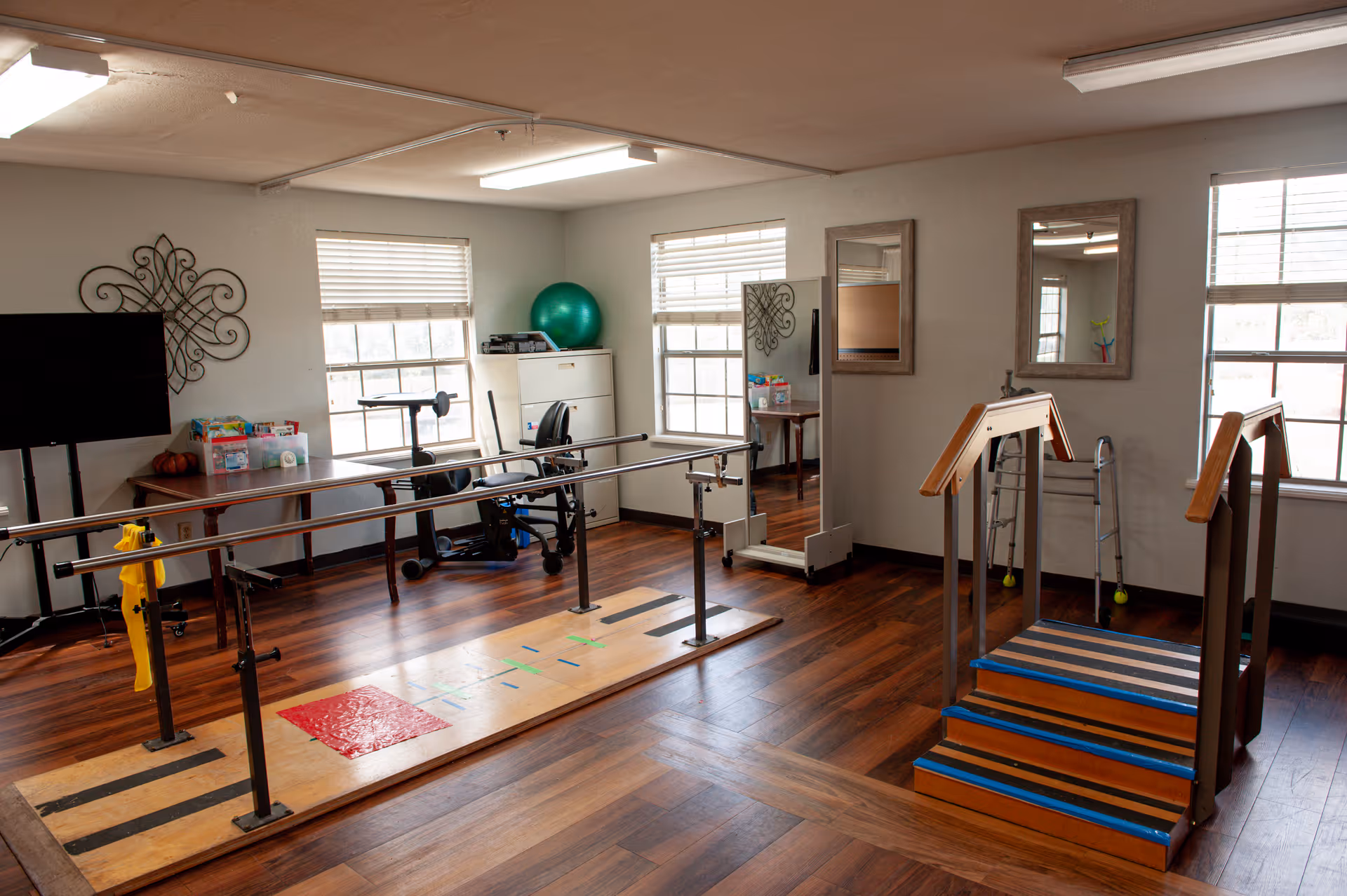 A rehabilitation room with wooden flooring featuring parallel bars for walking exercises and a small set of stairs with handrails for physical therapy. The room has three windows with blinds, a walker, a green exercise ball, a TV on a stand, a desk with supplies, a chair, a filing cabinet, and two mirrors mounted on the wall.