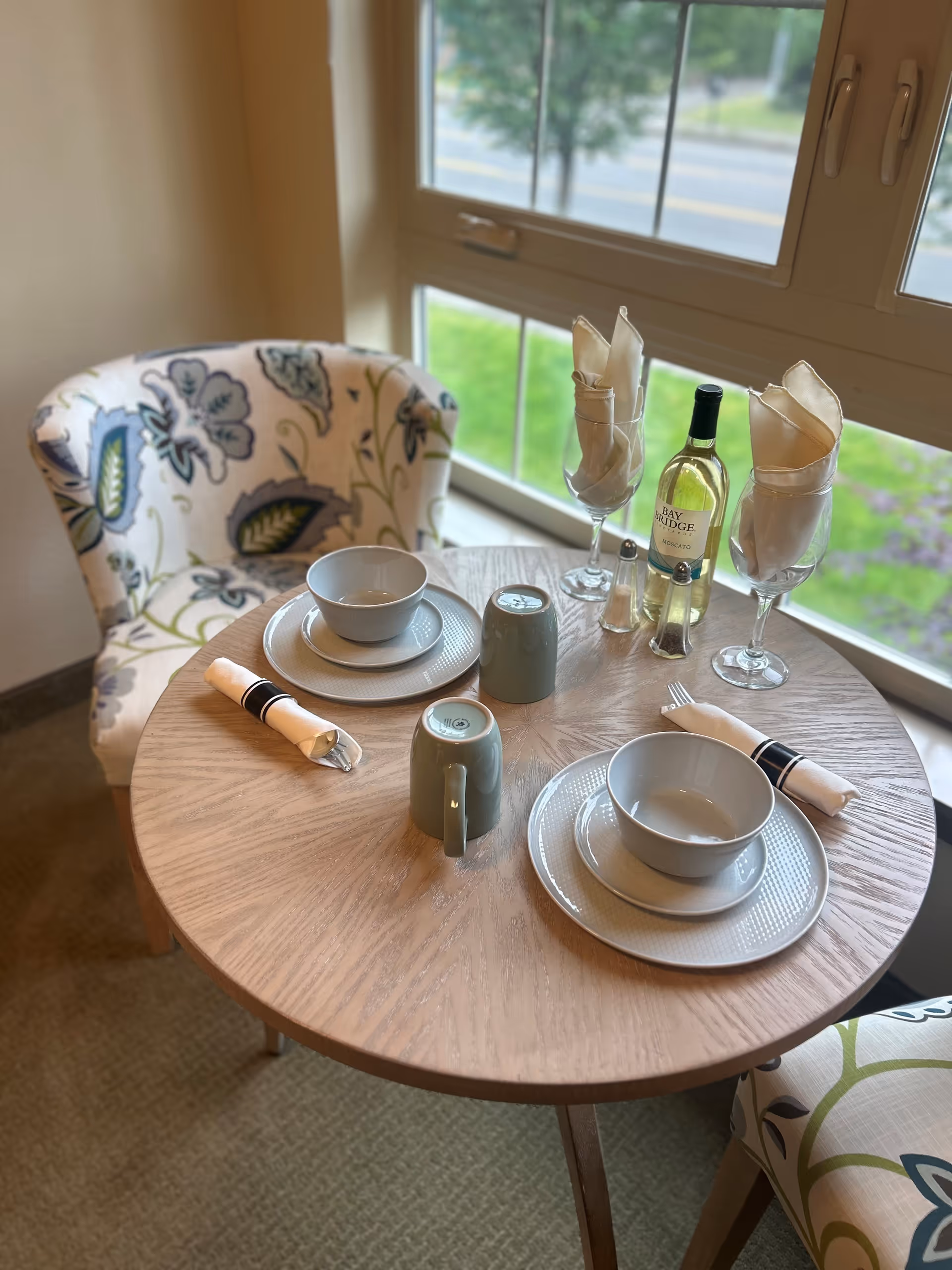A small round wooden dining table set for two with white plates, bowls, green mugs, and rolled napkins in napkin rings. Two floral-patterned upholstered chairs are positioned around the table. There is a bottle of Bay Bridge Moscato wine, two wine glasses with napkins inside, and a salt and pepper shaker on the table. A large window with a view of greenery and a road is in the background.