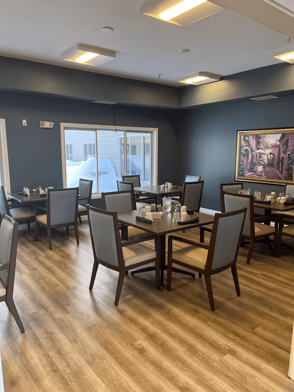 A dining room with several square wooden tables and cushioned chairs set for meals, wood-look flooring, and a large window showing snow outside.