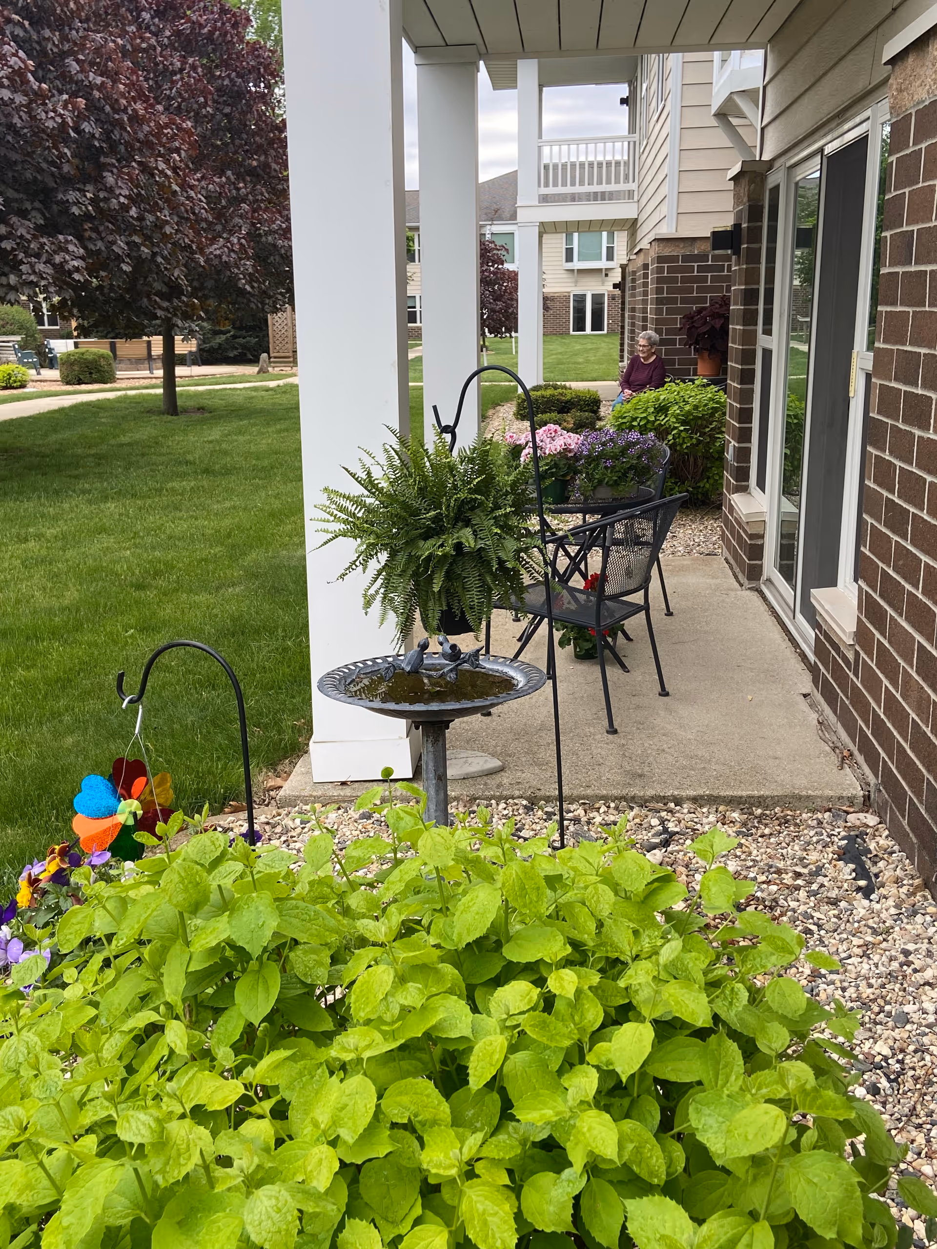 Outdoor patio area at The Meadows Senior Community featuring a small garden with green plants and flowers, a birdbath with a hanging fern, metal chairs and a table with potted flowers, and a senior woman sitting on a bench near the building. The area is surrounded by grass, trees, and other buildings.