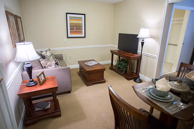 Small furnished living room with a sofa, coffee table, television on a stand, side tables with lamps, and a small dining table set.