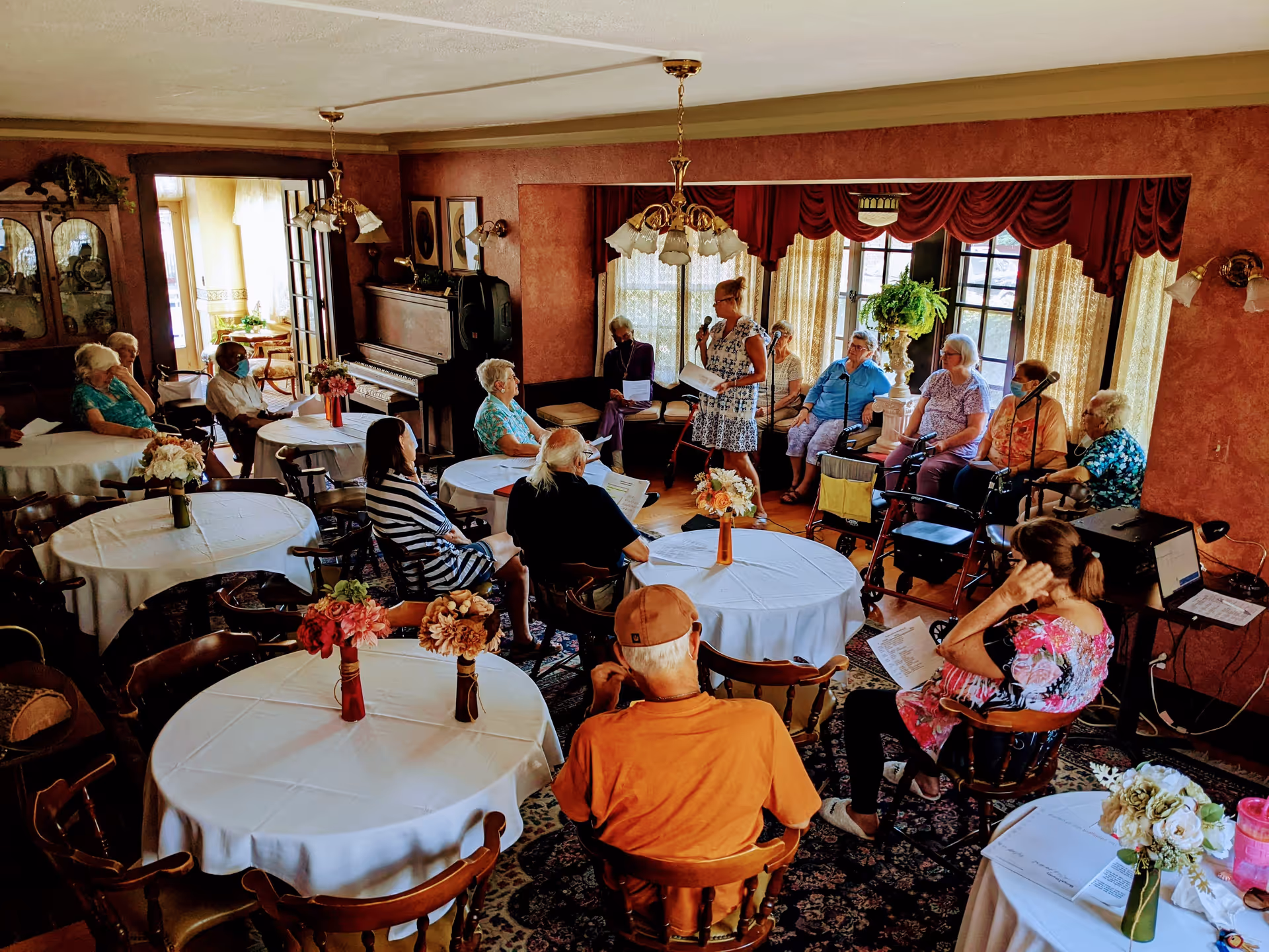 A group of elderly people seated around tables with white tablecloths in a warmly decorated room with red walls and large windows covered with curtains. A woman stands speaking into a microphone near a piano, while others listen attentively. The room has floral centerpieces on the tables and a cozy, communal atmosphere.